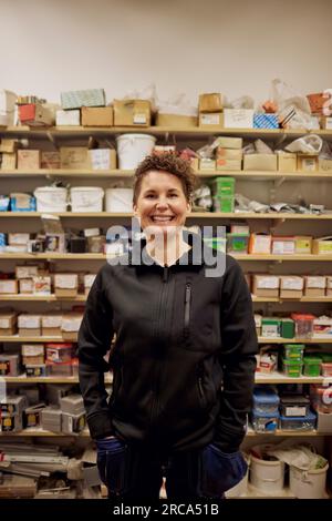 Portrait of happy mature female worker standing in front of rack at hardware store Stock Photo