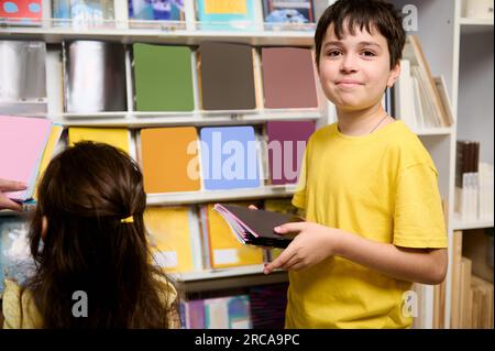 Caucasian happy kids, a teenage boy and a preschool girl choosing copybooks on shelves in school stationery store. Adorable primary school student boy Stock Photo
