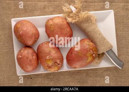 Several raw pink potatoes with white ceramic plate, jute napkin and vegetable peeler, macro, on jute cloth, top view. Stock Photo