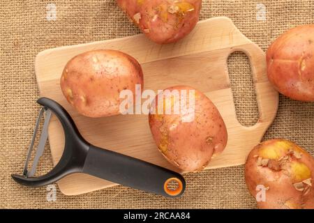 Several raw pink potatoes with wooden kitchen board and vegetable peeler knife, macro, on jute cloth, top view. Stock Photo