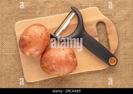 Several raw pink potatoes with wooden kitchen board and vegetable peeler knife, macro, on jute cloth, top view. Stock Photo