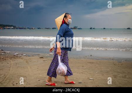 A middle-aged Vietnamese woman wearing a traditional conical hat carries a bag of seafood on My Khe beach, Danang, Vietnam. Stock Photo