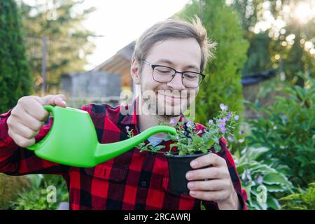 Young funny man florist in glasses holds a green watering can and watering plant outdoors Stock Photo