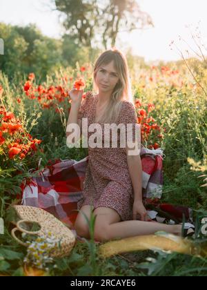 Pretty woman on plaid outdoor with blooming poppy flowers with sunset tones. Lady in dress and handbag with field flowers Stock Photo