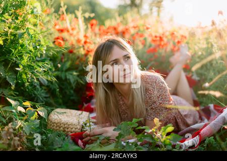 Pretty woman on plaid outdoor with blooming poppy flowers with sunset tones. Lady in dress and handbag with field flowers Stock Photo