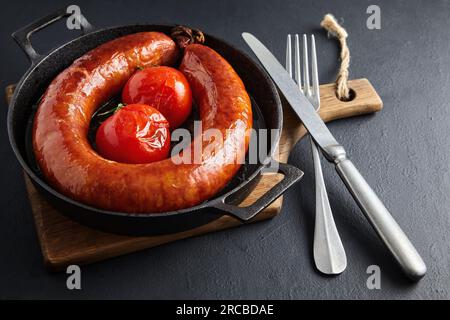 Ring of fried pork sausage and grilled tomatoes on black cast iron pan and fork with knife on wooden board on black stone table. Stock Photo