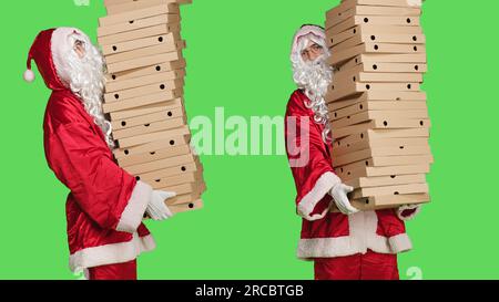Santa claus posing with pile of pizza boxes, carrying huge stack of delivery food, man wearing traditional character suit with white beard and glasses. Person in costume over greenscreen. Stock Photo