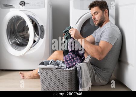 Man with clothes near washing machine. Laundry cleaning. Housework, homework, male housekeeper. Husband man doing laundry at home. Washer and dryer at Stock Photo
