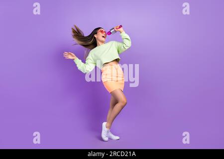 Full size photo of adorable pretty woman dressed green shirt stylish skirt sing in microphone on stage isolated on purple color background Stock Photo