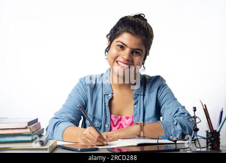 A pretty young Indian college student studying and looking at camera on study table and white background Stock Photo