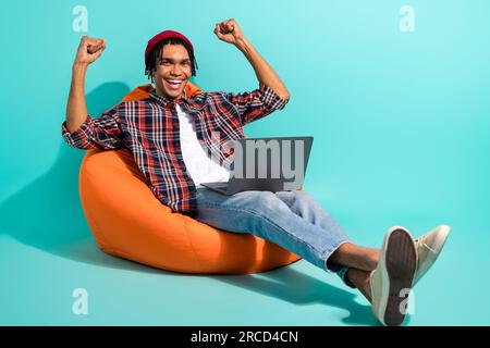 Full length photo of ecstatic guy wear hat sit on bean bag with laptop raising fists up win bet isolated on turquoise color background Stock Photo