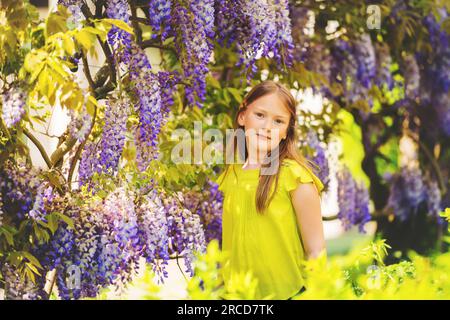 Outdoor stylish portrait of a cute little girl of 8-9 years old, standing next to beautiful purple wisteria flowers, wearing green blouse Stock Photo
