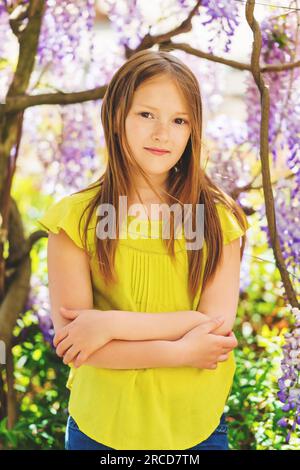 Outdoor stylish portrait of a cute little girl of 8-9 years old, standing next to beautiful purple wisteria flowers, wearing green blouse Stock Photo