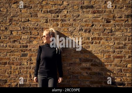 Attractive smiling blonde standing against brick wall Stock Photo