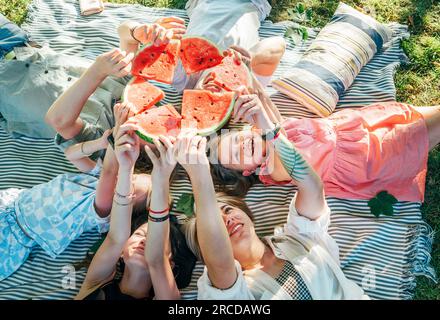 Young daughters with parents family lying on picnic blanket during weekend sunny day, smiling, laughing and rose up red juicy watermelon pieces. Famil Stock Photo