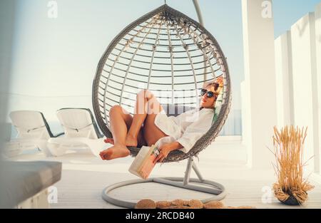 Beautiful middle-aged woman reading and lying in comfortable hanging chair on the open house terrace with bestseller book and enjoying the sunset sun Stock Photo