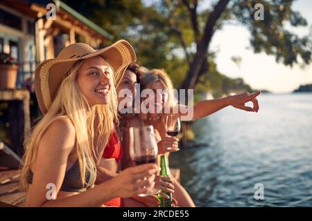 A group of female friends in bikini sitting in the bar on the river and enjoying the view and a drink on a beautiful summer day. Summer, river, vacati Stock Photo