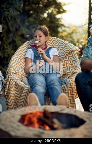 Lovely little girl eating grilled marshmallows, sitting outdoors by a fireplace in a cozy chair, enjoying summer day in the yard. Lifestyle concept. Stock Photo