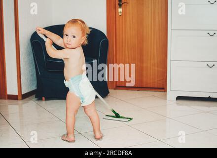 Adorable 1 year old baby boy helping with cleaning, looking back over the shoulder, wearing diaper, holding a mop Stock Photo