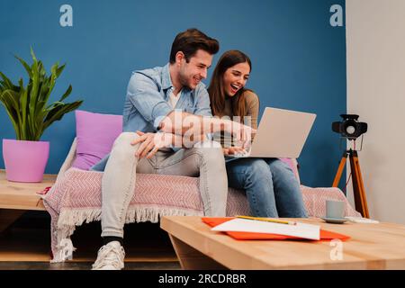 Happy young couple watching the laptop screen, checking finances, planning budget or mortgage sitting at couch of their apartment. Smiling husband showing on his wife's notebook a online shopping sale. High quality photo Stock Photo