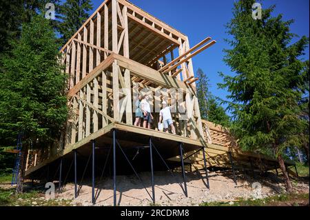 Man engineer showing to couple process of constructing wooden framed house. Youthful pair of investors inspecting their future dwelling in mountains near forest. Concept of contemporary construction. Stock Photo