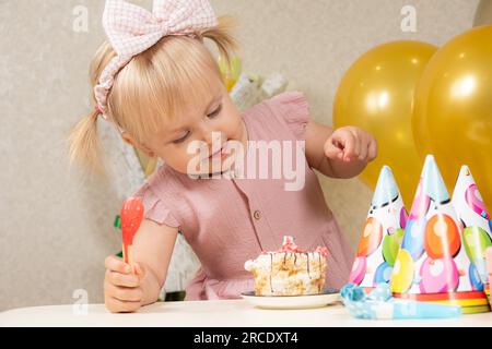 Little blonde girl with two ponytails and a bow on her head eating cake with a spoon and hands on her birthday Stock Photo