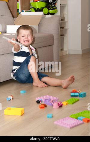 A small Caucasian beautiful boy was upset and crying while sitting on the floor with scattered toys. He does not want to remove the children's multi-c Stock Photo