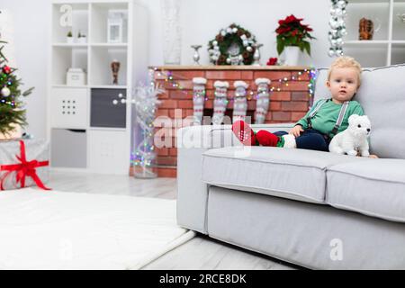 A little boy in a Christmas outfit sits on a couch against a fireplace decorated with lights. Stock Photo