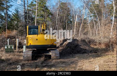 Yellow hydraulic backhoe excavator with tracks is parked near utility boxes at a construction site, with trees in the distance. Stock Photo