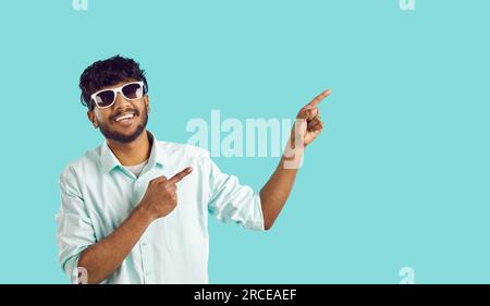 Young positive Indian man in sunglasses points with finger of two hands at copy space Stock Photo