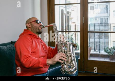 young latin man saxophonist of venezuelan ethnicity with beard and glasses is at home sitting on the sofa inspired playing music, music concept, copy Stock Photo