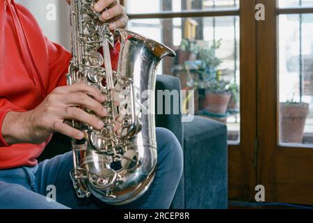 closeup in profile with copy space of caucasian male hands of unrecognizable man sitting on the sofa playing a saxophone inside home. Stock Photo