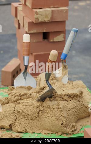 Three builders building trowels in pile of cement mortar, with stack of bricks behind. Stock Photo