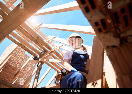 Woman standing on ladder, checking readiness of wooden roof frame Stock Photo