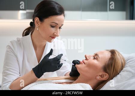 Brunette doctor makes rejuvenating injection in the chin of client Stock Photo