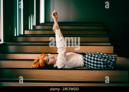 A stunning red-haired Asian schoolgirl, donned in her uniform, lies on her desk in a moment of recess, stretching her arms up. Beauty and youth. Stock Photo