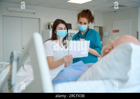 Female doctors with piece of paper visiting male patient in ward Stock Photo