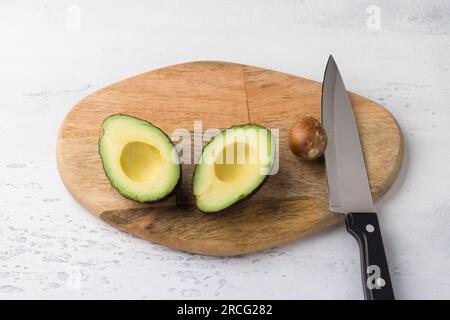 Fresh avocado cut in half and avocado pit with a knife on a wooden board on a light gray background, top view. Stock Photo