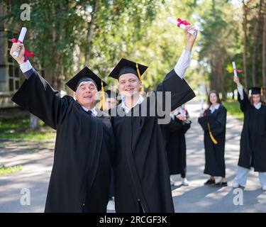 A group of graduates in robes outdoors. An elderly man and a young guy congratulate each other on receiving a diploma. Stock Photo