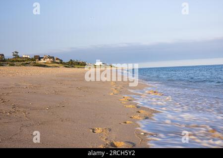 A landscape scenery of waves rolling onto the beach, houses in the background, seashells and footprints in the sand, love heart written in sand. Stock Photo