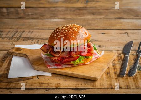 Homemade burger with bacon, lettuce, tomato, eggs and avocado on wooden cutting board. Side view, close up Stock Photo