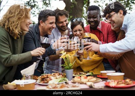 Adults around a set table, toasting with red wine, under decorative lights and twilit trees Stock Photo