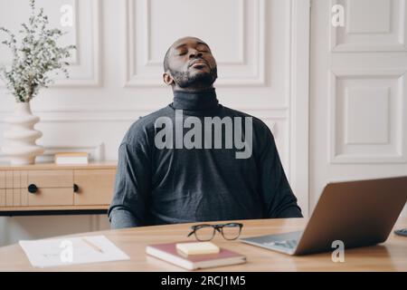 Tired Afro American employee rests, removes glasses, closes eyes, and leans back, recovering from remote work. Stock Photo