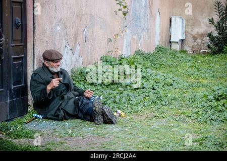 old man maybe homeless sat on the ground outside a doorway in Rome wearing a flat cap and overcoat and with legs outstretched showing his boot soles Stock Photo
