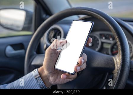 Detail of the hand of a driver holding a mobile phone with a blank screen on the steering wheel of the car. Stock Photo