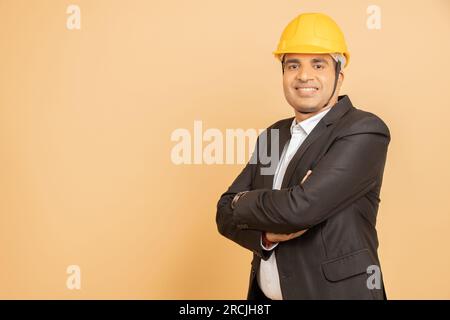 Happy indian engineer wearing black suit and yellow hard hat or helmet standing cross arms isolated over beige background. Copy space. Stock Photo