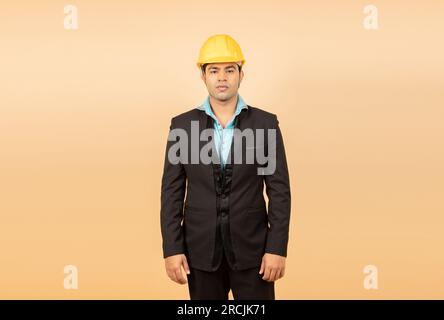 Young indian engineer or architect wearing black formal suit and yellow hard hat or helmet standing isolated over beige background. Industrial and con Stock Photo