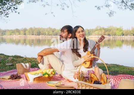 Young couple in love sitting back to back to playing ukulele guitar while picnic time together. Stock Photo