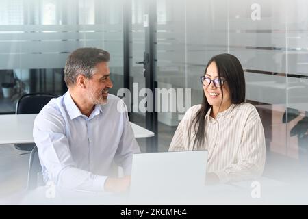 Two happy multicultural professionals working in office using laptop, talking. Stock Photo