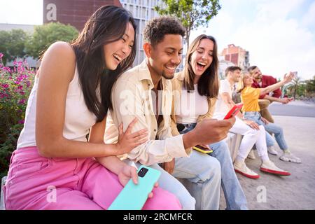 Multiracial happy group of friends using phone and smiling together outdoor. People addicted mobile. Stock Photo